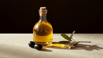 Editorial still-life of a glass bottle of golden extra virgin olive oil with whole olives and an olive branch sprig, styled on cream stone against a deep olive-brown background with a lime-green accent