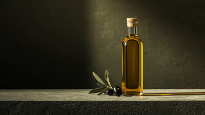 Editorial still-life of a glass bottle of golden olive oil with olives and an olive branch sprig, styled on textured stone in deep olive-brown, green and soft cream tones with a lime accent, no text