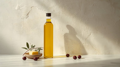 Editorial still-life of a glass bottle of golden extra virgin olive oil with olives and an olive branch sprig, styled with a small dish of butter on a cream stone surface, in deep olive and lime tones