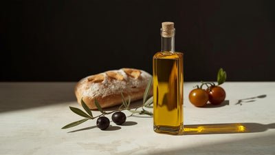 Editorial still-life of a glass bottle of golden olive oil with olives, an olive branch sprig, rustic bread and tomatoes, styled on cream stone in a deep olive-green and lime-accented palette