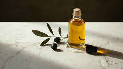 Editorial still-life of a glass bottle of golden olive oil with whole olives, an olive branch sprig and a small dropper, styled on a cream stone surface in dark olive, deep green and soft off-white tones with a touch of lime accent.