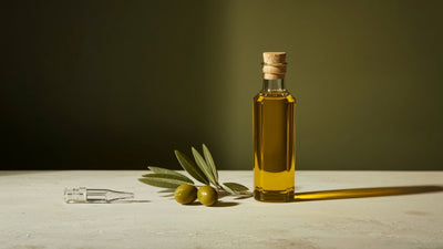 Editorial still-life of a glass bottle of golden extra virgin olive oil with whole olives, a small olive branch sprig and a glass dropper, styled on a cream stone surface against a deep dark olive-brown background with a soft lime-green accent