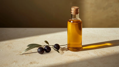 Editorial still-life photograph of a glass bottle of golden olive oil with whole olives, an olive branch sprig, and a small glass dropper, styled on textured cream stone in deep olive-brown and lime-green tones.
